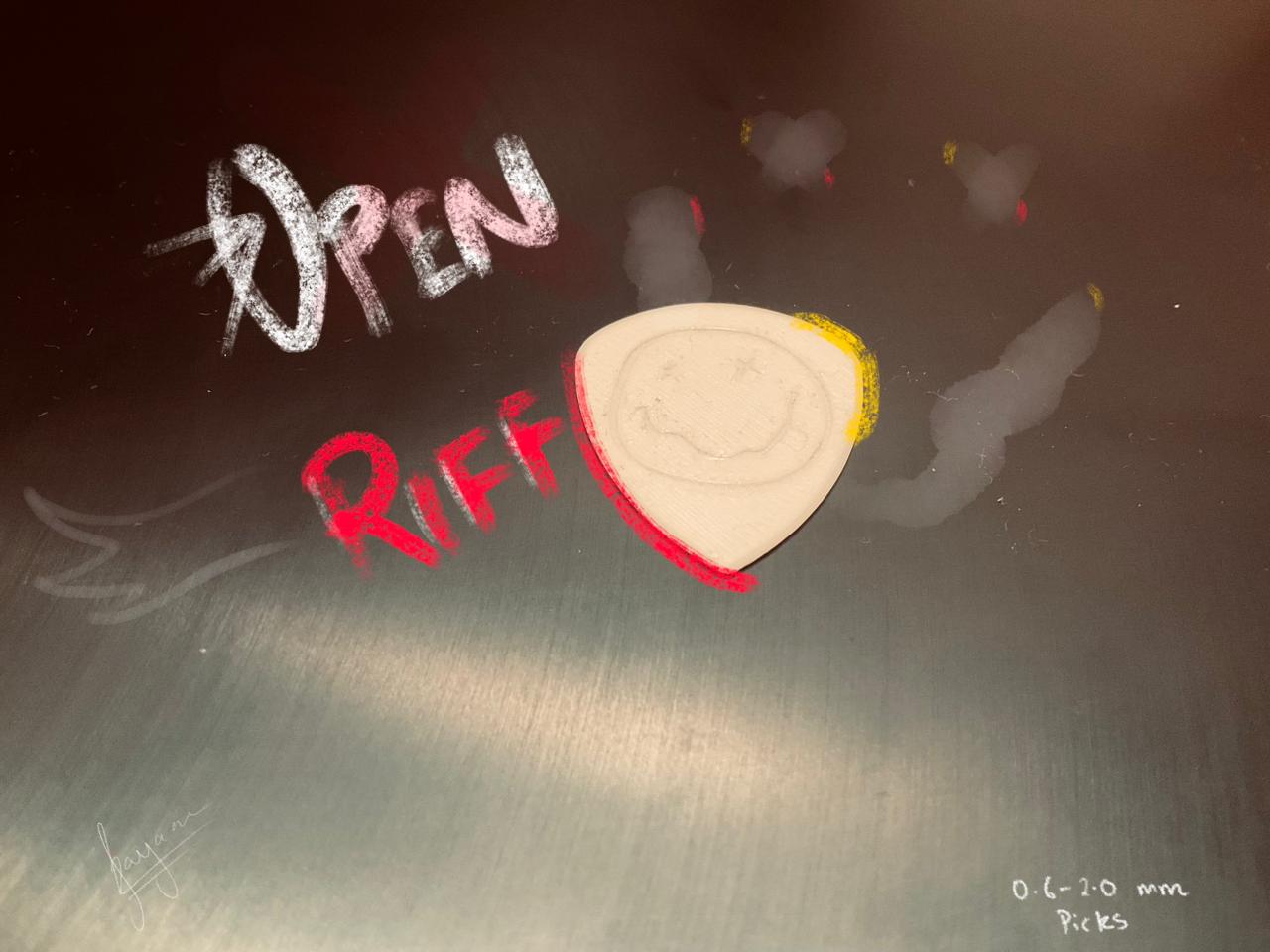 A bone-white 3D-printed pick with a smiley-face engraving on a brushed-metal bench, with 'OPEN RIFF' sketched in chalk beside it.